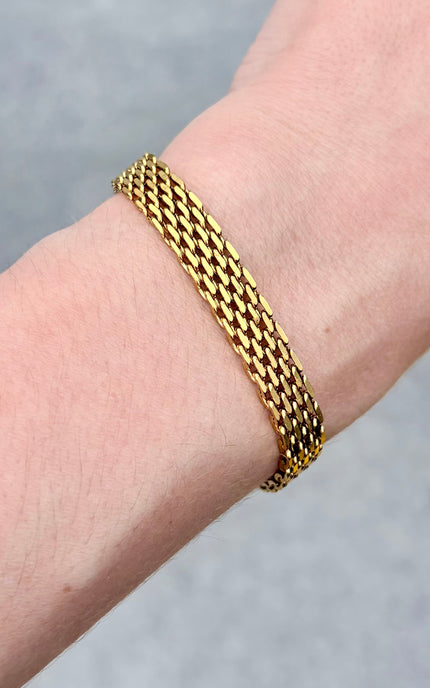 A close-up of a gold-plated three-strand chain bracelet worn on a person's wrist against a soft gray background.