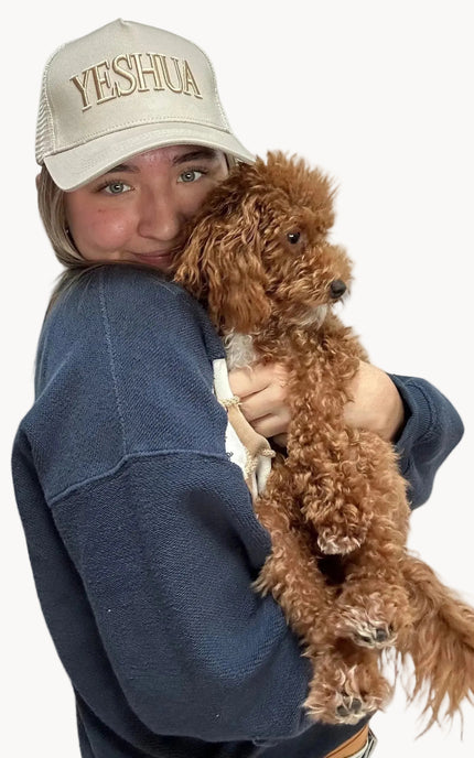 Person holding a brown dog wearing a cap with 'YESHUA' text on a white background