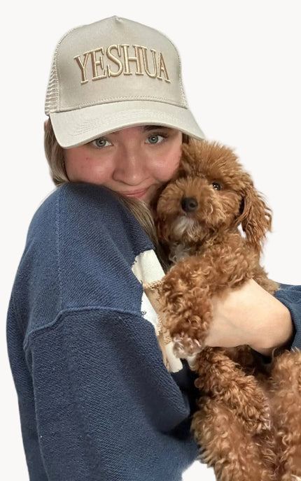 Person wearing a cap with 'YESHUA' text, holding a small brown dog on a white background