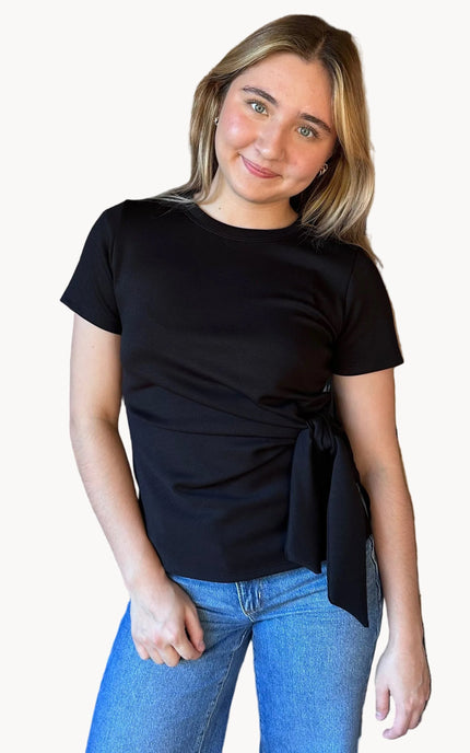 Woman wearing a black top with a tie detail and blue jeans on a beige background