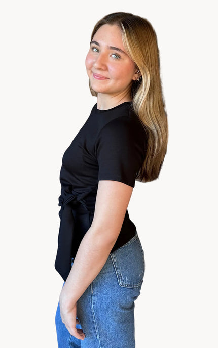 Woman in black top and blue jeans standing against a brick wall.