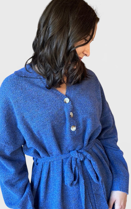 A woman wearing a blue knit belted cardigan dress with a buttoned neckline, shown from the side against a plain light gray background.