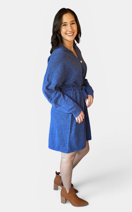 A woman modeling a blue textured long-sleeve belted knit dress against a light gray studio background.