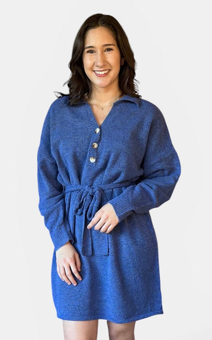 A woman wearing a blue knit belted dress with a buttoned collar, standing against a plain light gray studio background.