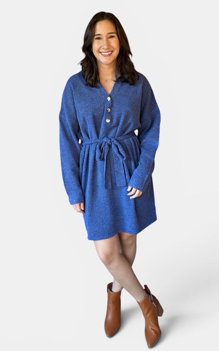 A woman modeling a blue knit sweater dress with a buttoned neckline, long sleeves, a matching tie belt, and brown ankle boots against a light gray studio background.