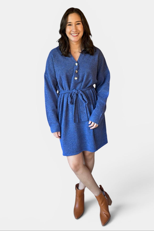 A woman modeling a blue knit sweater dress with a buttoned neckline, long sleeves, a matching tie belt, and brown ankle boots against a light gray studio background.