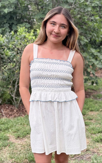 Front view of a feminine romantic white cotton smocked mini dress with a square neckline and flared skirt on a model with hands in pockets.