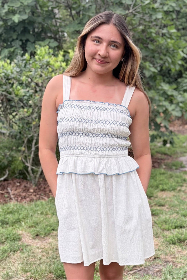 Front view of a feminine romantic white cotton smocked mini dress with a square neckline and flared skirt on a model with hands in pockets.