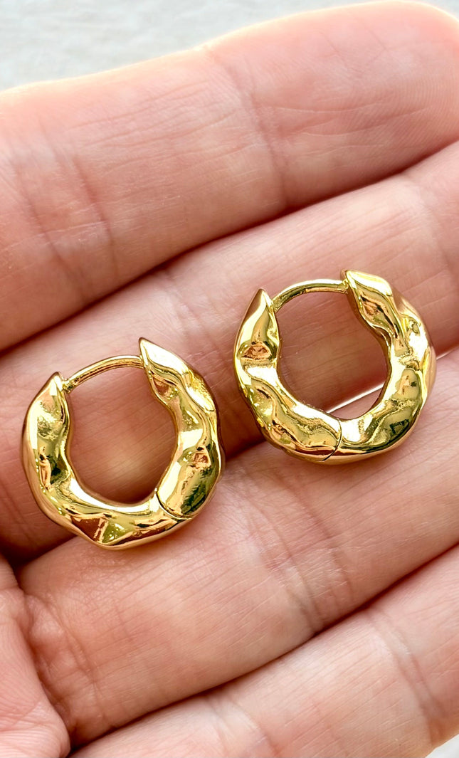 Gold hoop earrings held between two fingers on a neutral background