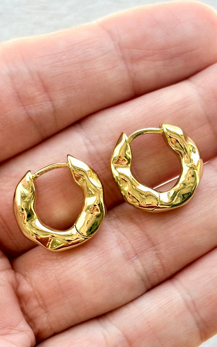 Gold hoop earrings held between two fingers on a neutral background