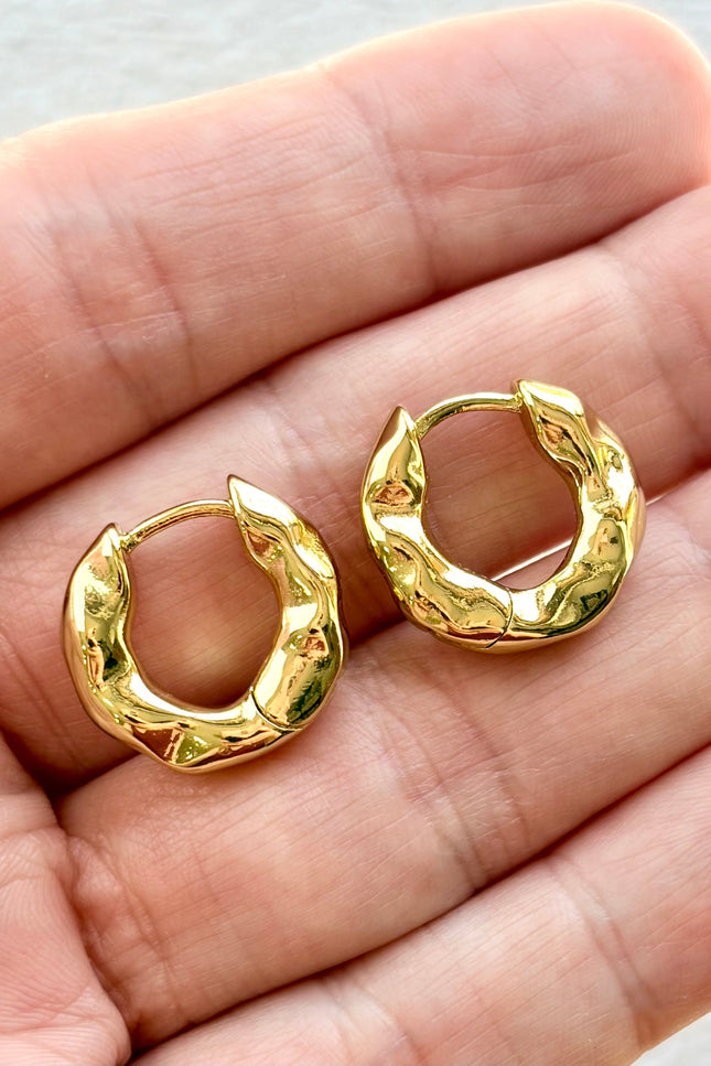 Gold hoop earrings held between two fingers on a neutral background