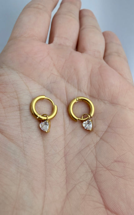 Gold hoop earrings with heart crystal drop held in hand showing scale, sparkle detail, and 18k gold PVD finish
