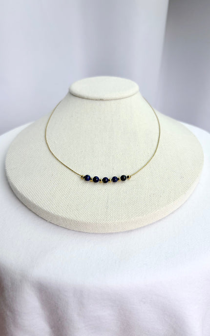 A deep blue lapis lazuli beaded necklace beautifully draped on a minimalist linen necklace display stand. The shot captures the vibrant contrast of the natural blue gemstone beads and the rich texture of the polished 18k gold PVD twisted chain in warm, natural lighting.