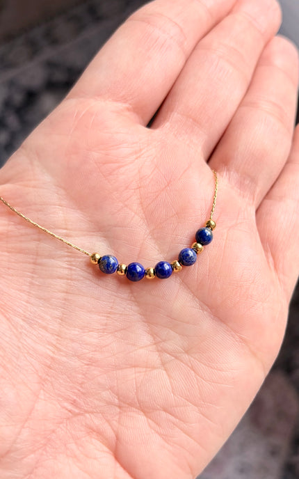 A handheld close-up of a dainty gold necklace resting in the palm of a hand. The shot highlights the rich, deep blue of the natural lapis lazuli beads alternating with polished gold accents on a delicate twisted chain.