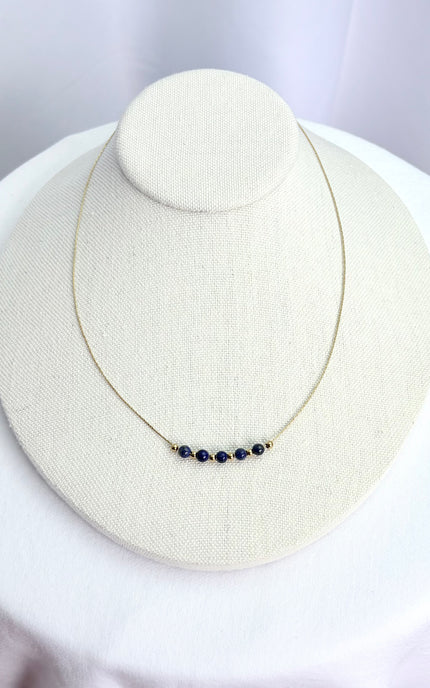 A deep blue lapis lazuli beaded necklace beautifully draped on a minimalist linen necklace display stand. The shot captures the vibrant contrast of the natural blue gemstone beads and the rich texture of the polished 18k gold PVD twisted chain in warm, natural lighting.