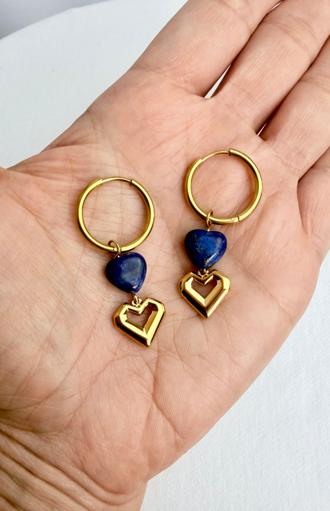 A handheld close-up of a pair of gold heart hoop earrings resting in the palm of a hand. The shot highlights the rich, deep blue of the natural lapis lazuli heart stones stacked beautifully above the polished open gold heart charms.