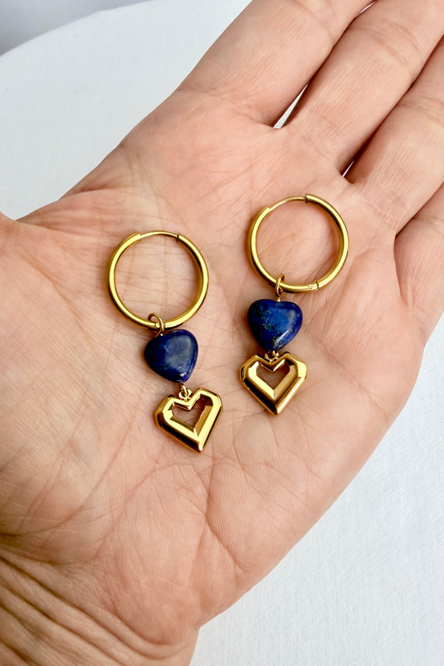 A handheld close-up of a pair of gold heart hoop earrings resting in the palm of a hand. The shot highlights the rich, deep blue of the natural lapis lazuli heart stones stacked beautifully above the polished open gold heart charms.