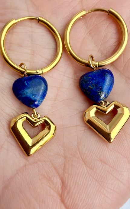A handheld close-up of a pair of gold heart hoop earrings resting in the palm of a hand. 