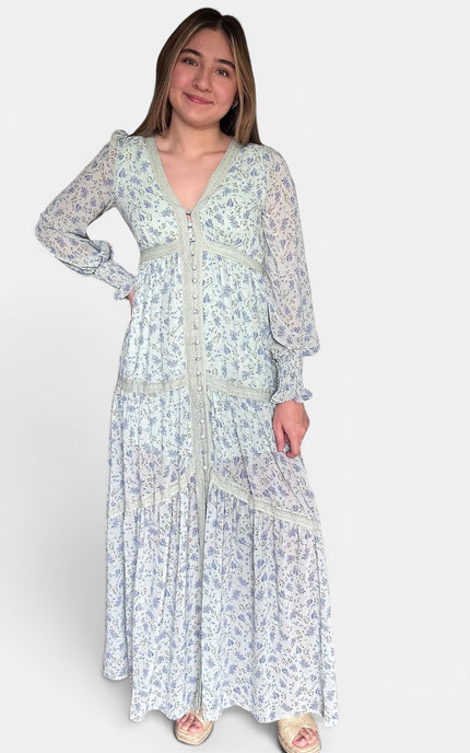 A woman modeling a long-sleeve maxi dress with a light blue and white floral pattern, lace trim, and a button-up front, set against a plain light gray background.