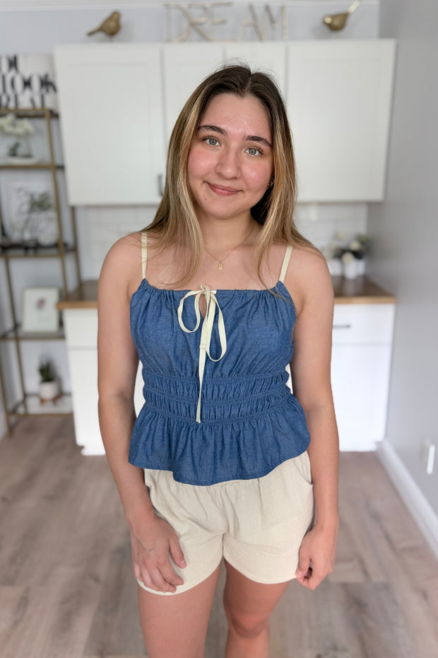 Soft cotton-blend denim cami with a fitted shirred bodice, adjustable front tie, and flowy peplum hem, styled for an easy, feminine summer look