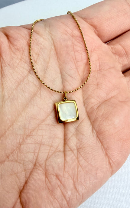 Close-up shot of an 18k gold PVD plated square pendant necklace held in a palm, showcasing the genuine white sea shell inlay, polished gold bezel, and delicate 9.2mm scale of the piece in natural light.