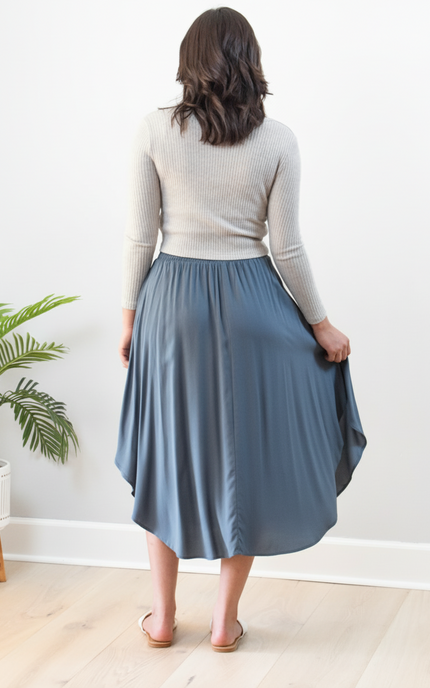 Person wearing a blue skirt and beige top in a room with a plant and small table.