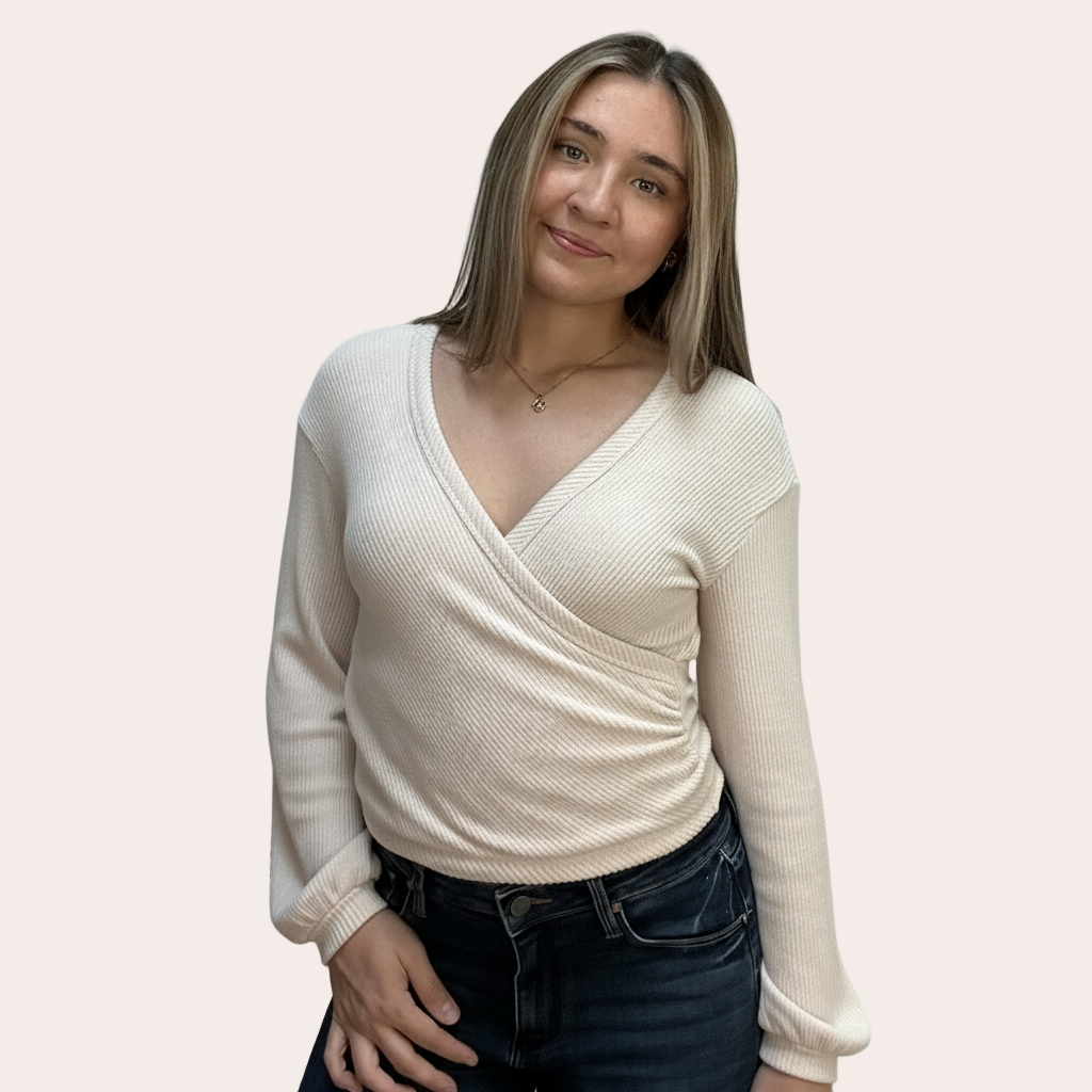 Woman wearing a cream wrap sweater and dark jeans on a light pink background