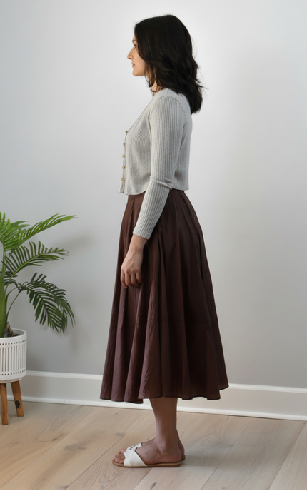 Woman wearing a white sweater and brown skirt standing in a room with a plant and wooden floor.